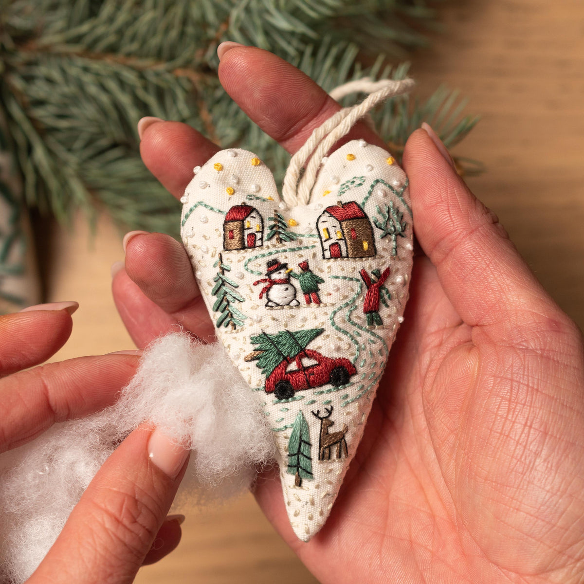 May include: A white heart-shaped ornament with red and green embroidery depicting a winter scene with houses, a snowman, a car, and a deer. The ornament is being held in a hand and is being stuffed with white fiberfill.