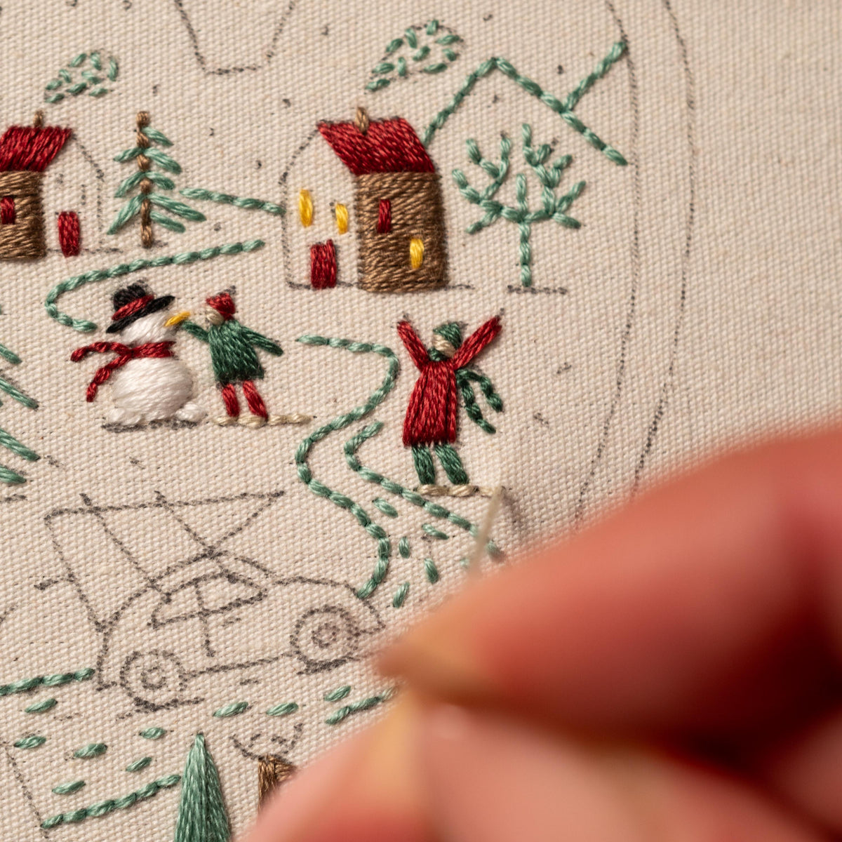 May include: A close-up of a hand embroidering a winter scene on a white canvas. The scene includes a snowman, two people, a house, and a car. The embroidery is done in green, red, and brown thread.