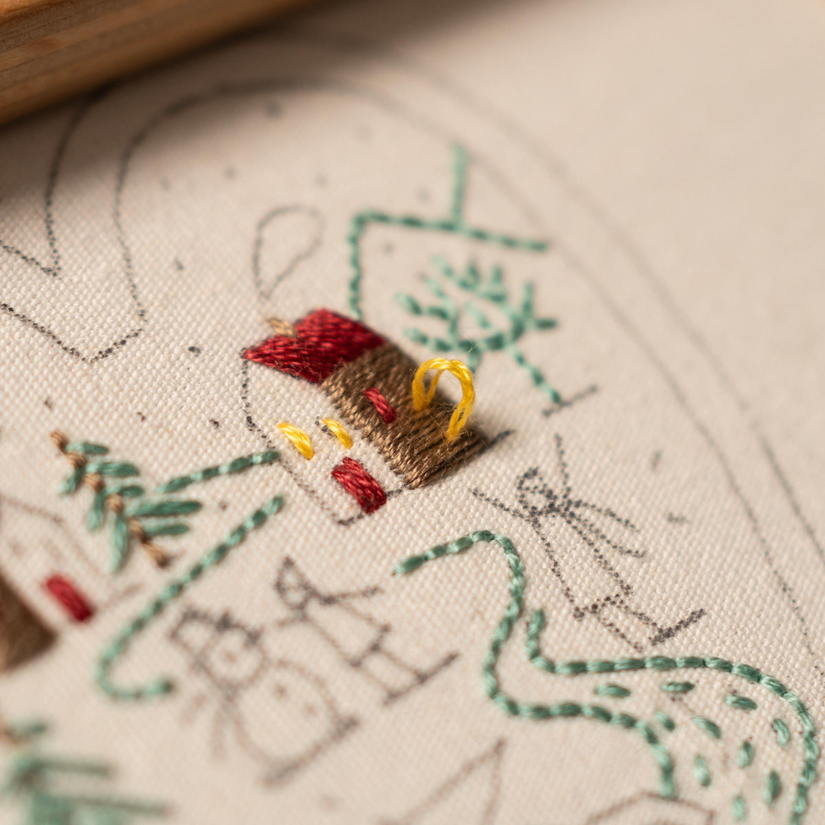 May include: Close-up of a hand-stitched embroidery design on a cream-colored fabric. The design features a small brown house with a red roof and a yellow door, surrounded by green trees and a winding path.
