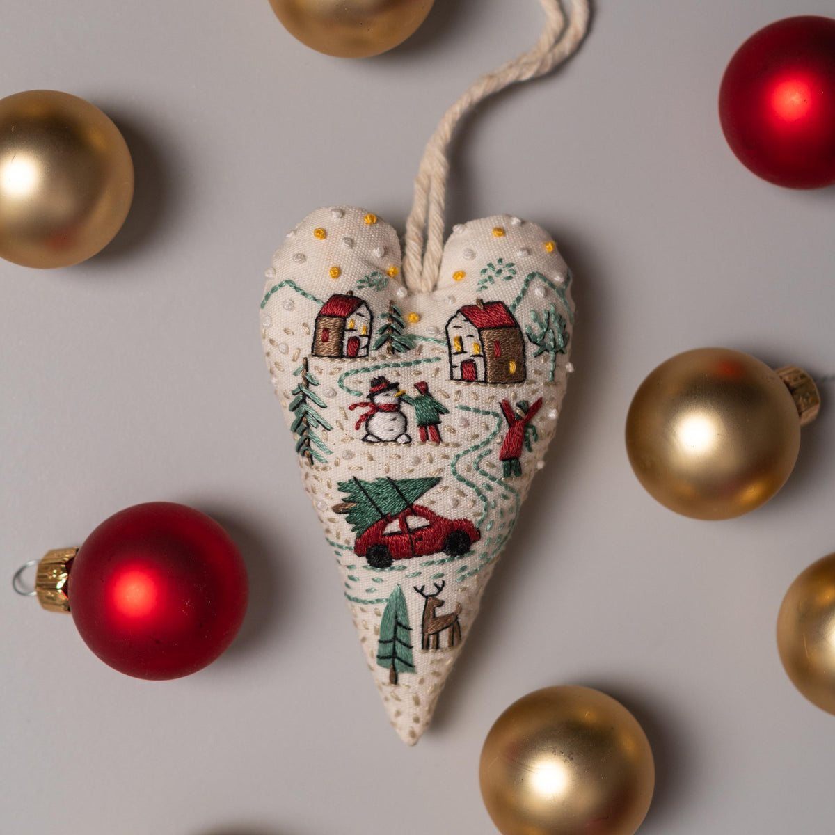 May include: A white heart-shaped ornament with a winter scene embroidered on it. The scene includes houses, a snowman, a car with a Christmas tree on top, and a deer. The ornament is surrounded by gold and red Christmas ornaments.