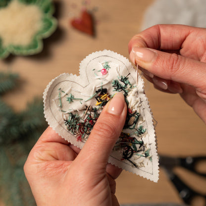 May include: A white heart-shaped fabric with a colorful embroidered design is being sewn shut with a needle and thread.