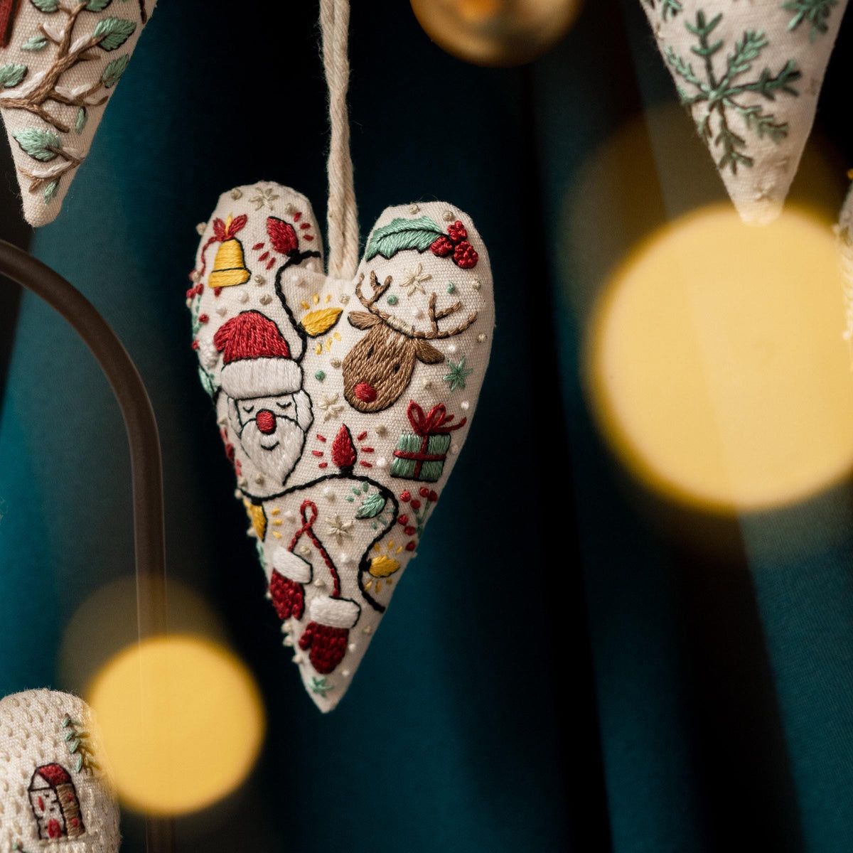 May include: A white heart-shaped ornament with colorful embroidered Christmas designs, including Santa Claus, a reindeer, and a bell. The ornament is hanging from a string and is surrounded by blurry lights.