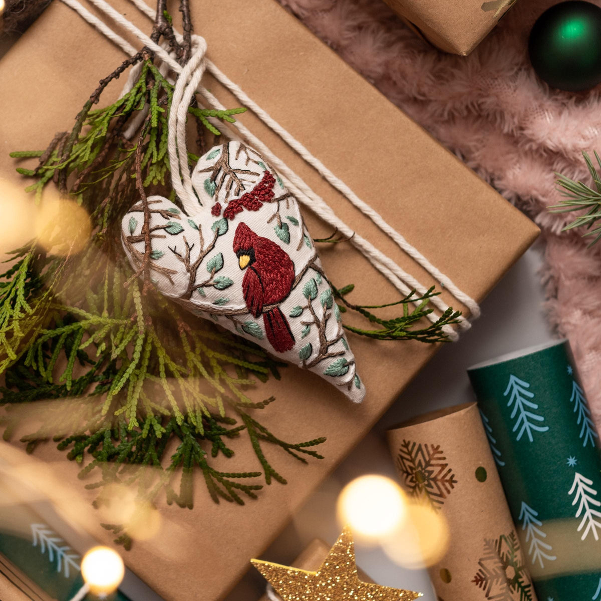 May include: A white heart-shaped ornament with a red cardinal embroidered on it. The ornament is hanging from a brown gift box wrapped with twine. The ornament is surrounded by green pine branches.