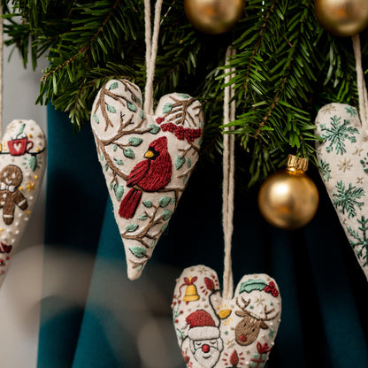 May include: A set of five heart-shaped Christmas ornaments with embroidered designs. The ornaments feature a cardinal, a gingerbread man, Santa Claus, a reindeer, and snowflakes. The ornaments are hanging from a Christmas tree.