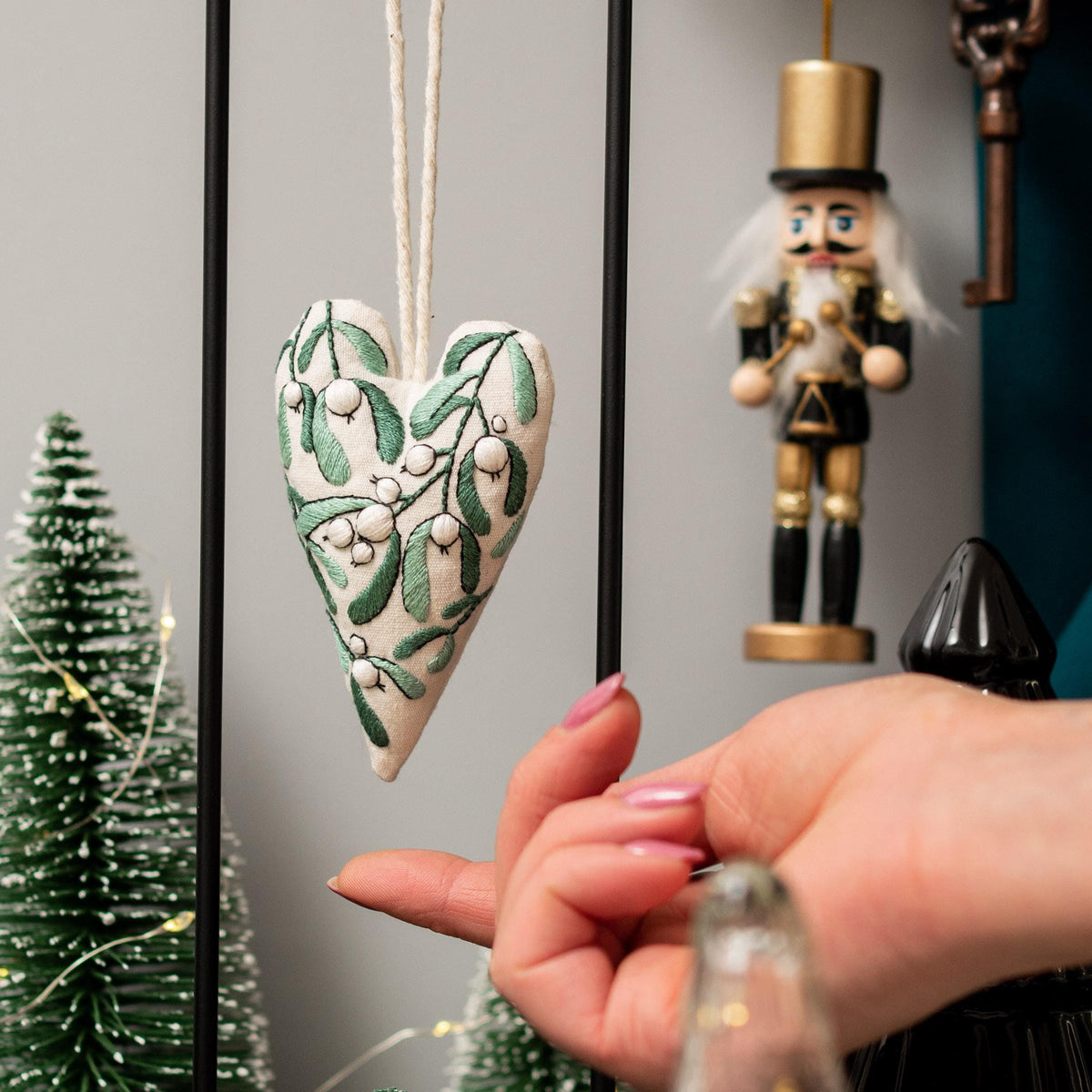 May include: A white heart-shaped ornament with green and white embroidery of mistletoe. The ornament is hanging from a white string and is in focus. The background is out of focus and includes a wooden nutcracker and a small green Christmas tree.