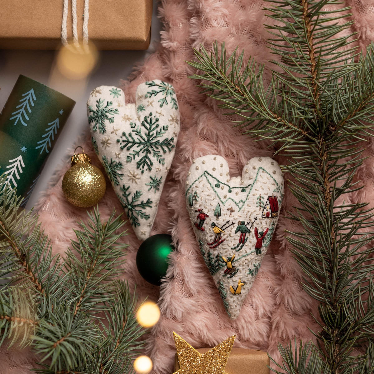 May include: Two white heart-shaped ornaments with embroidered designs. One ornament features a snowflake pattern in green and white, while the other depicts a winter scene with skiers and a small house. The ornaments are on a pink fuzzy surface with