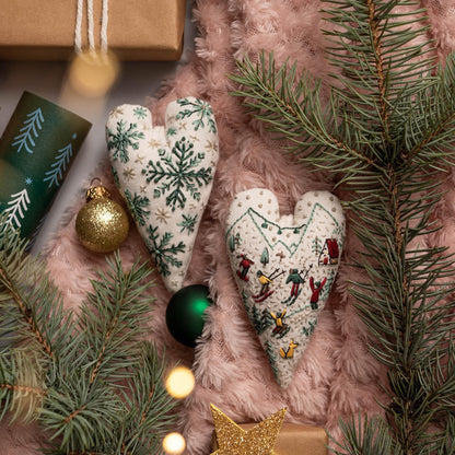 May include: Two white heart-shaped ornaments with embroidered designs. One ornament features a snowflake pattern in green and white, while the other depicts a winter scene with skiers and a small house. The ornaments are on a pink fuzzy surface with