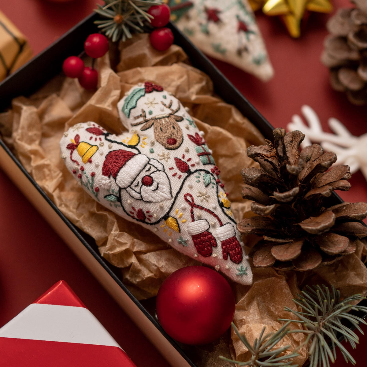 May include: A white heart-shaped ornament with red, green, and yellow embroidery depicting Santa Claus, a reindeer, and other Christmas symbols. The ornament is nestled in brown paper with a red Christmas ball and pine branches.