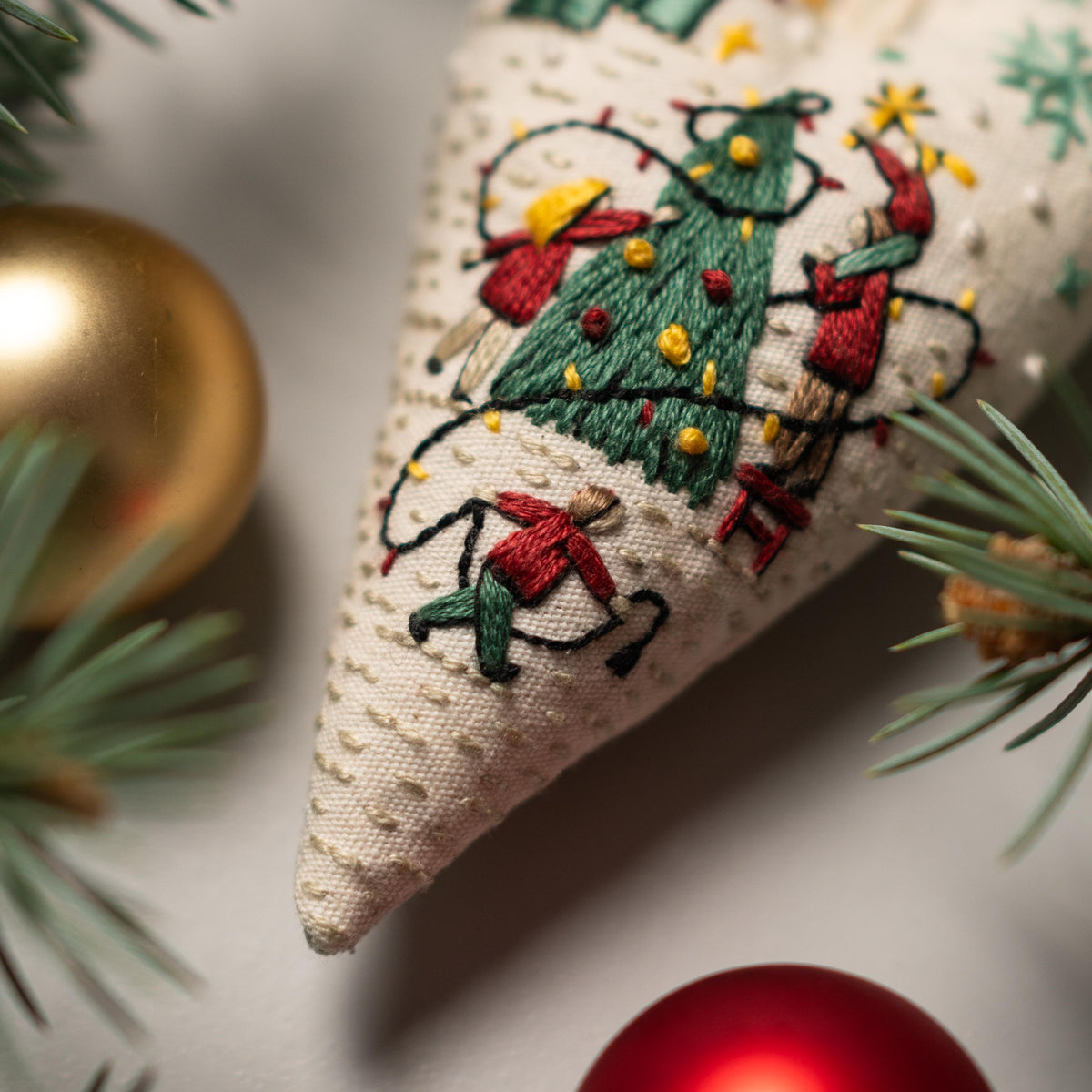 May include: A white fabric Christmas tree ornament with embroidered details of a green Christmas tree, red and yellow figures, and a string of lights. The ornament is decorated with a white background and a textured surface.