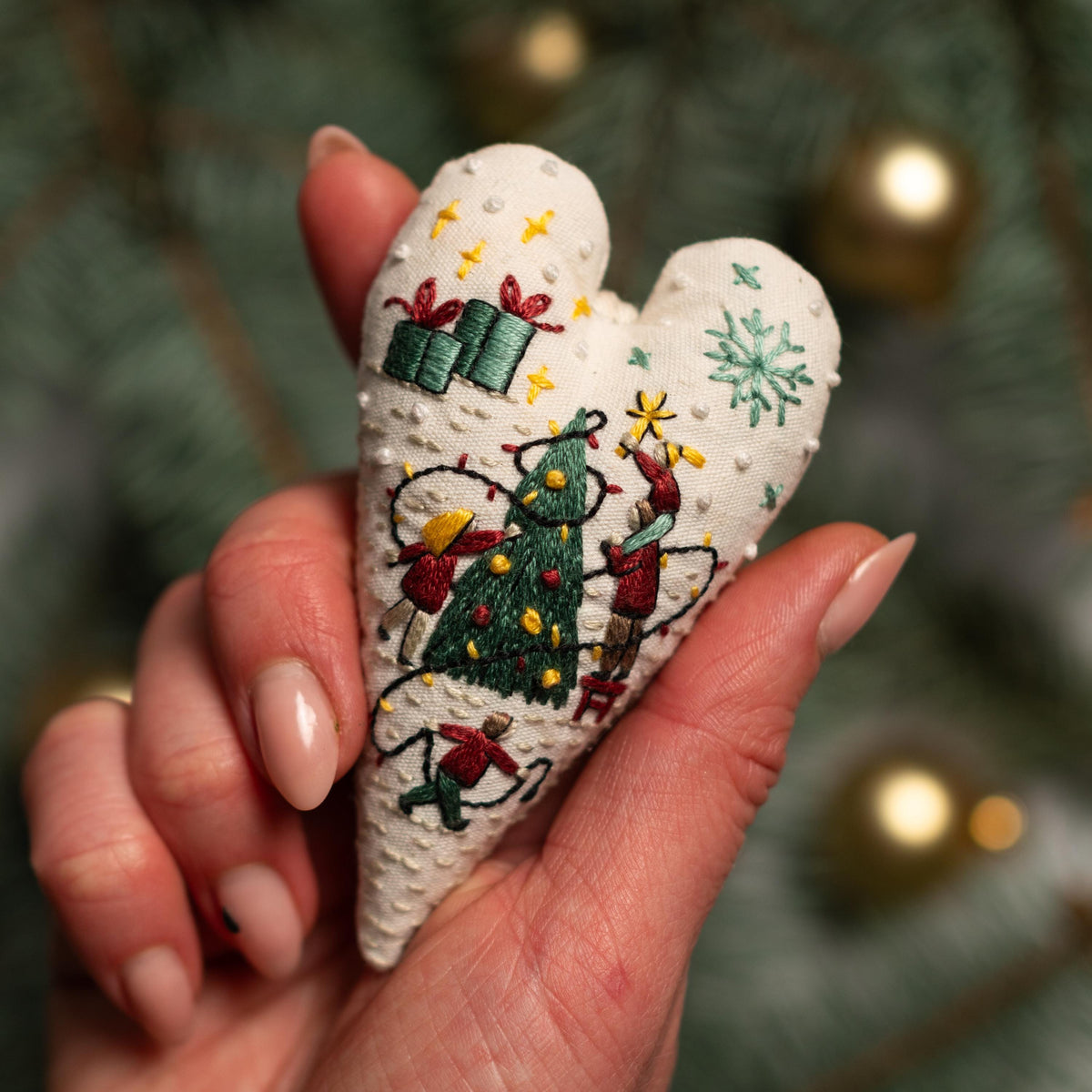 May include: A white heart-shaped fabric ornament with embroidered Christmas scenes. The ornament features a Christmas tree, presents, and people decorating the tree. The ornament is decorated with white dots.