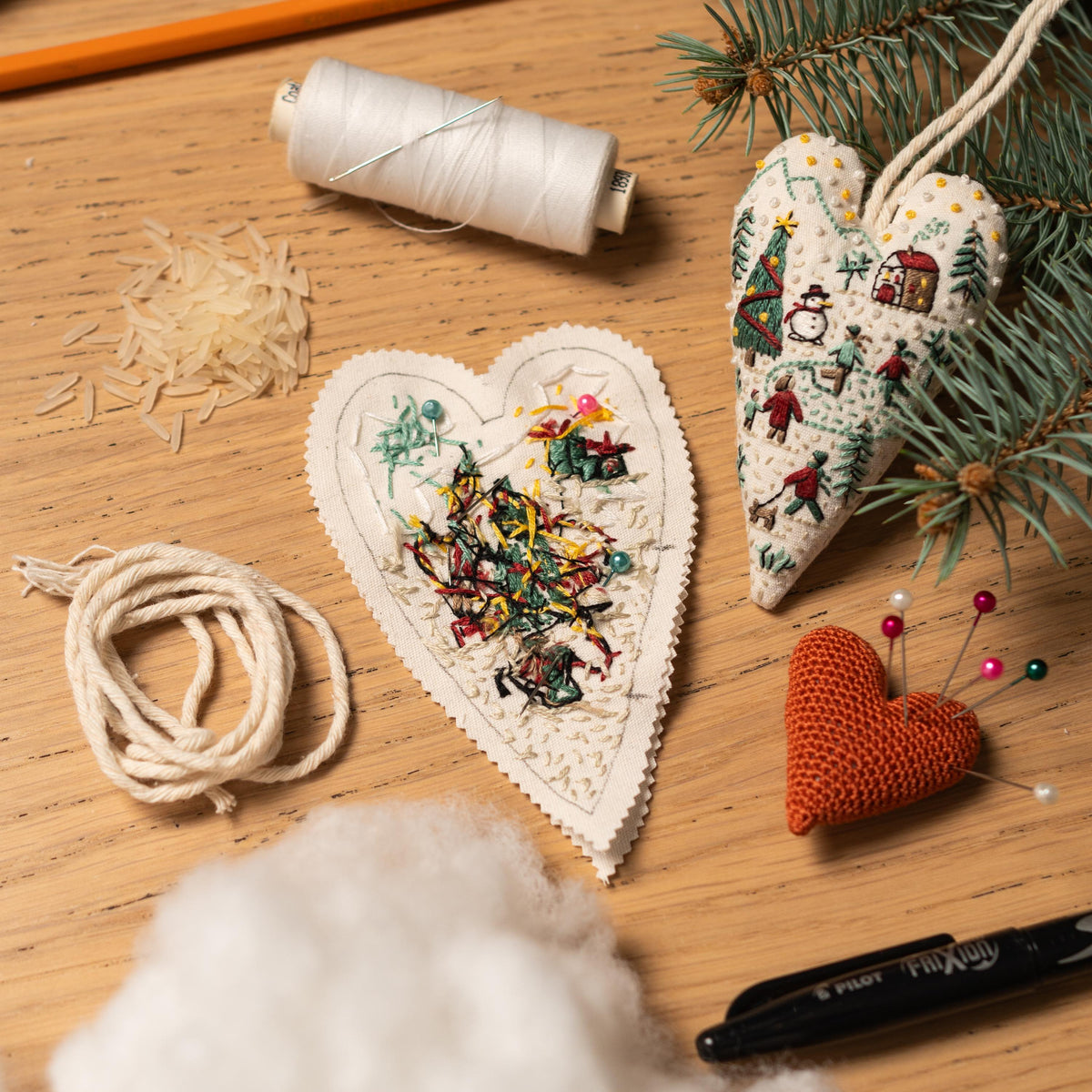 May include: Two white felt heart-shaped ornaments with colorful embroidery of Christmas trees and other holiday designs. The ornaments are surrounded by craft supplies including white thread, white stuffing, a spool of white thread, a pencil, and a 