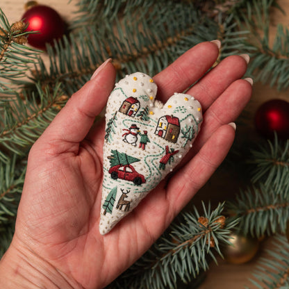 May include: A white heart-shaped ornament with embroidered details of a winter scene. The scene includes a red car, a snowman, a Christmas tree, and houses. The ornament is decorated with white beads.