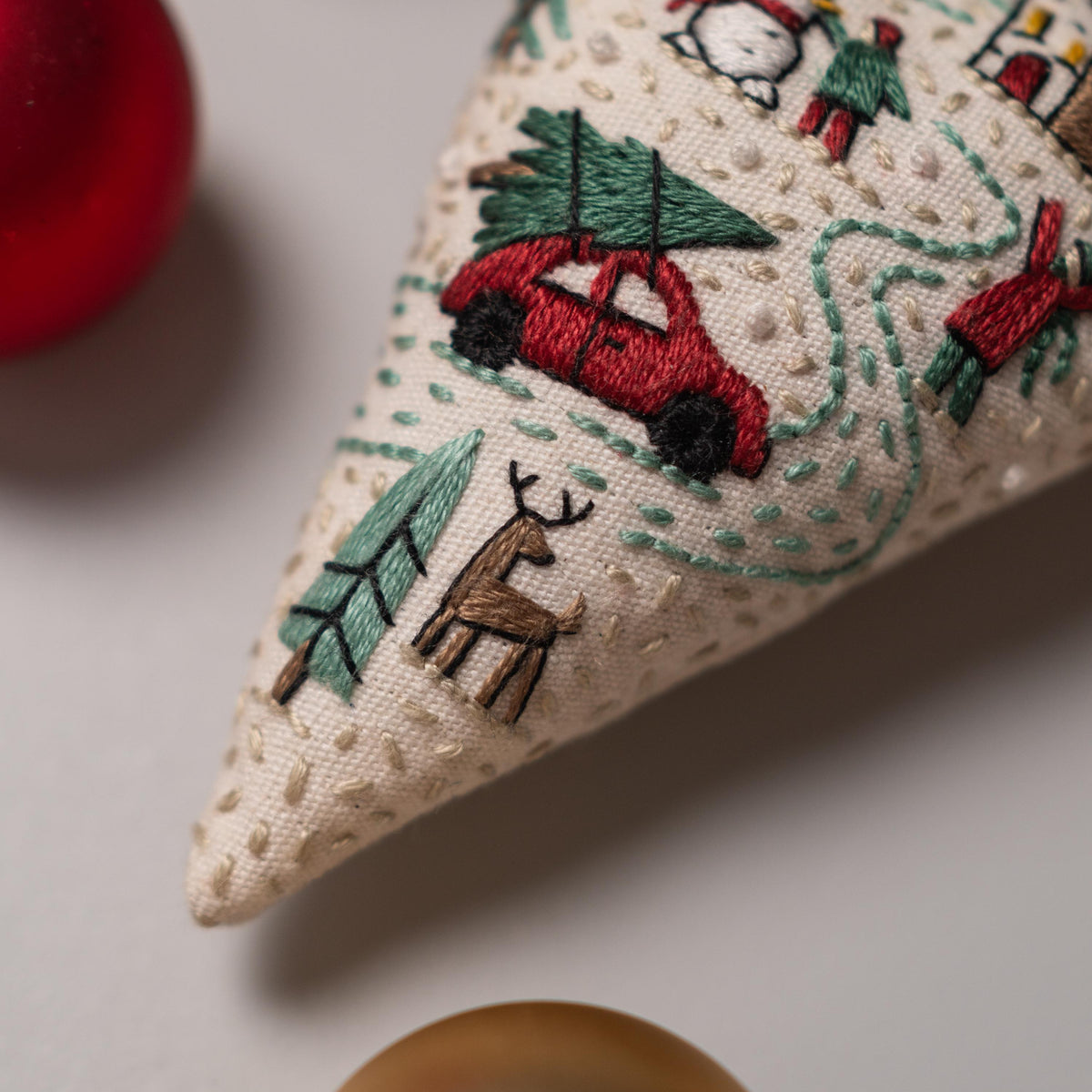 May include: A beige fabric cone with a red car carrying a green Christmas tree, a brown deer, and green trees embroidered on it.