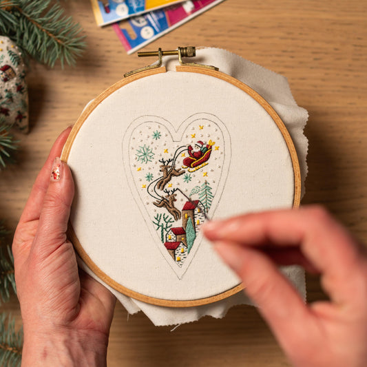 May include: A close-up of a hand embroidering a heart-shaped design on white fabric. The design features Santa Claus in a sleigh pulled by reindeer, flying over a small town with snow falling.
