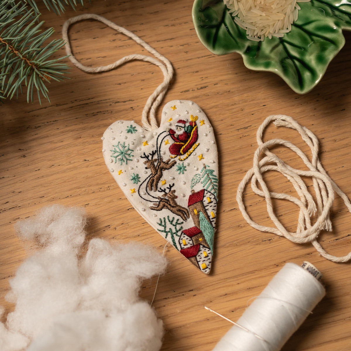 May include: A white heart-shaped ornament with a Christmas scene embroidered on it. Santa Claus is flying in a sleigh pulled by reindeer over a snowy village. The ornament is hanging from a white string.
