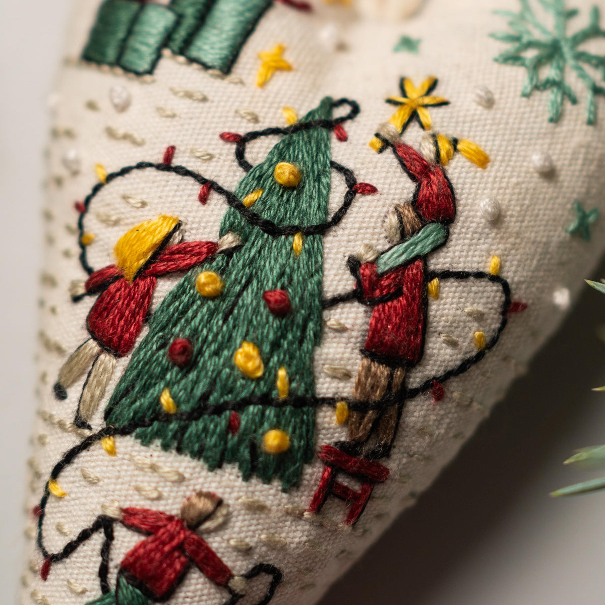 May include: Embroidered Christmas ornament featuring a green Christmas tree with yellow lights, red ornaments, and two people decorating it. One person is standing on a red stool and the other is holding a star.