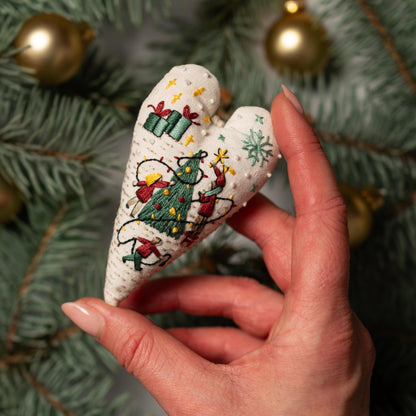 May include: A white heart-shaped ornament with a Christmas scene embroidered on it. The scene includes a green Christmas tree, red and white figures, and yellow lights.