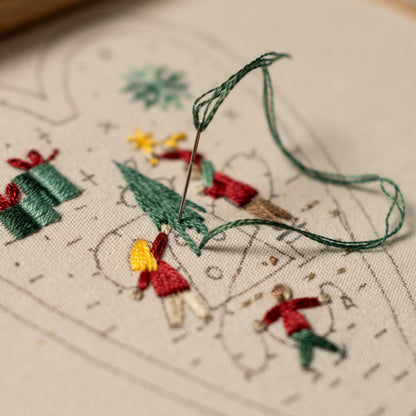 May include: Close-up of a needle and green thread being used to embroider a Christmas scene on white fabric. The scene includes a Christmas tree, presents, and people.