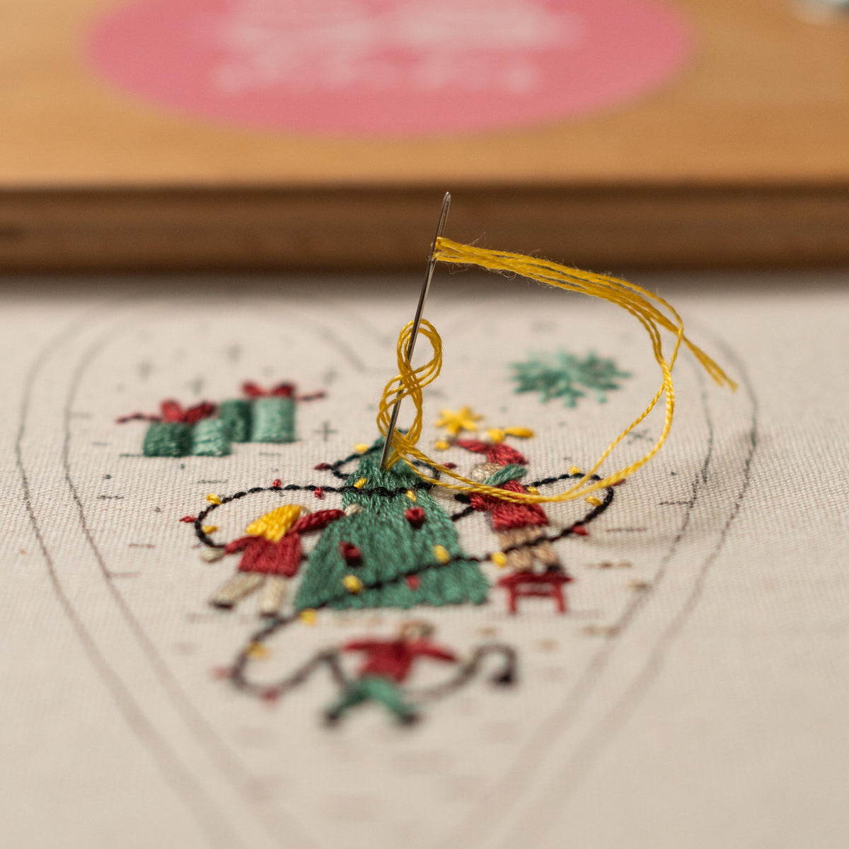 May include: Close-up of a needle and yellow thread being used to embroider a heart-shaped design on white fabric. The design features a Christmas tree, presents, and people.