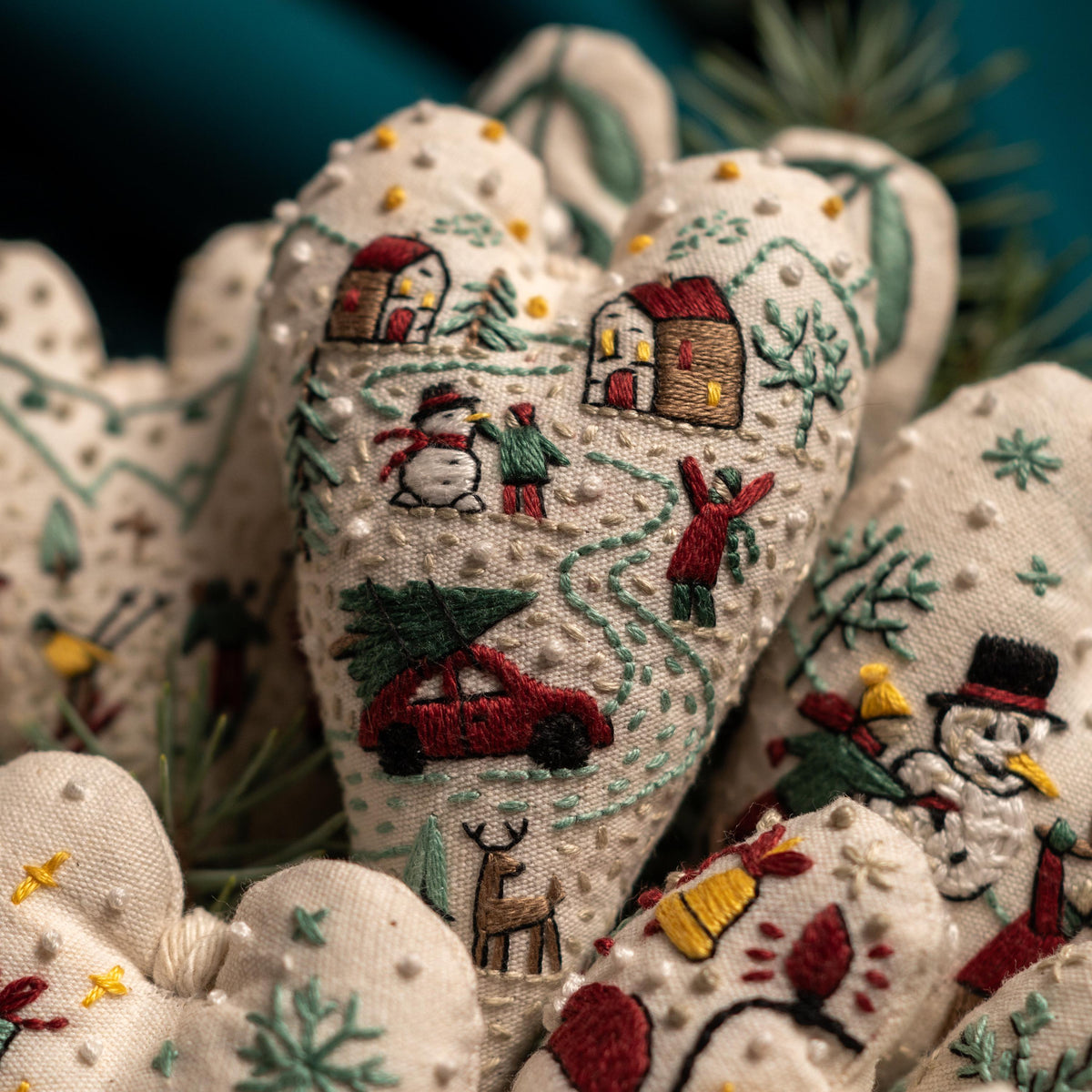 May include: A collection of handmade heart-shaped ornaments with embroidered winter scenes. The ornaments feature a red car carrying a Christmas tree, snowmen, houses, and other wintery details. The ornaments are made of white fabric with green, red