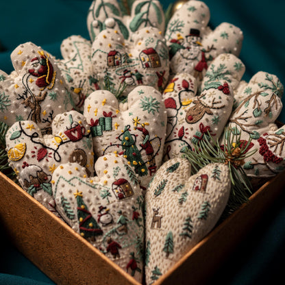 May include: A box filled with handmade heart-shaped ornaments. Each ornament is made of white fabric and features a different embroidered Christmas scene, including a snowman, a reindeer, a Christmas tree, and a Santa Claus.