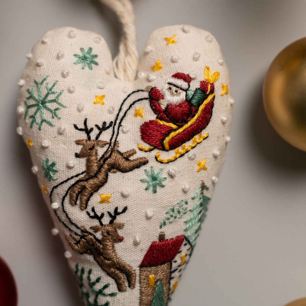 May include: A white heart-shaped ornament with a red and white Santa Claus in a sleigh pulled by two reindeer. The ornament is decorated with green and white snowflakes, yellow stars, and white dots.