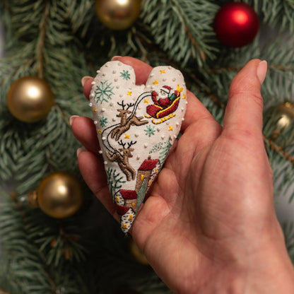 May include: A white heart-shaped ornament with a red, green, and white embroidered design of Santa Claus in a sleigh pulled by reindeer. The ornament is decorated with gold glitter and white snowflakes.