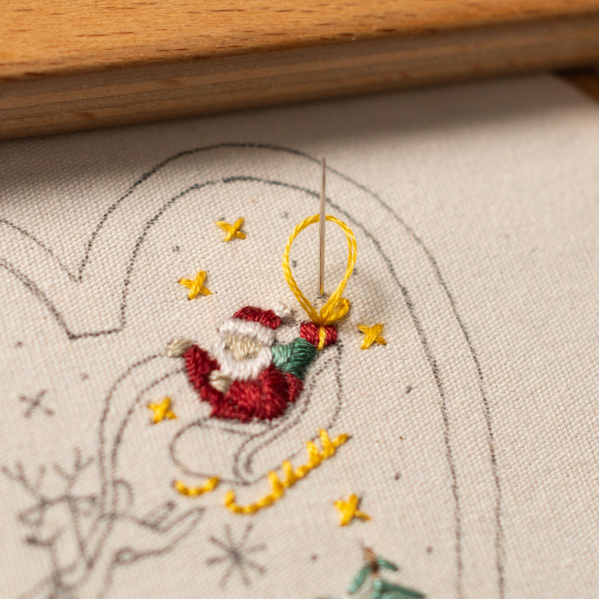May include: Close-up of a needle and thread being used to create a yellow bow on a white fabric with a Santa Claus design embroidered on it.