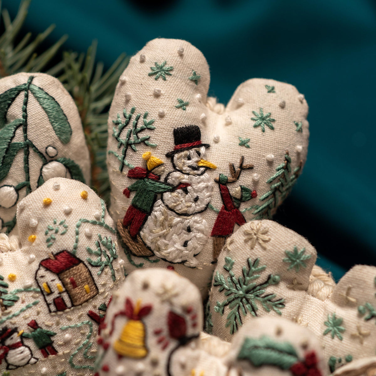 May include: A set of handmade heart-shaped ornaments with embroidered winter scenes. The ornaments feature a snowman, a house, and a bell. The ornaments are made of white fabric and are decorated with green, red, and white embroidery.