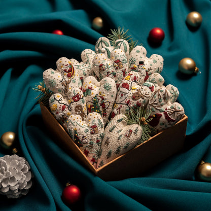 May include: A box filled with handmade felt heart ornaments decorated with winter holiday scenes. The ornaments are in various shades of white, red, green, and brown. The box is sitting on a teal fabric surface.