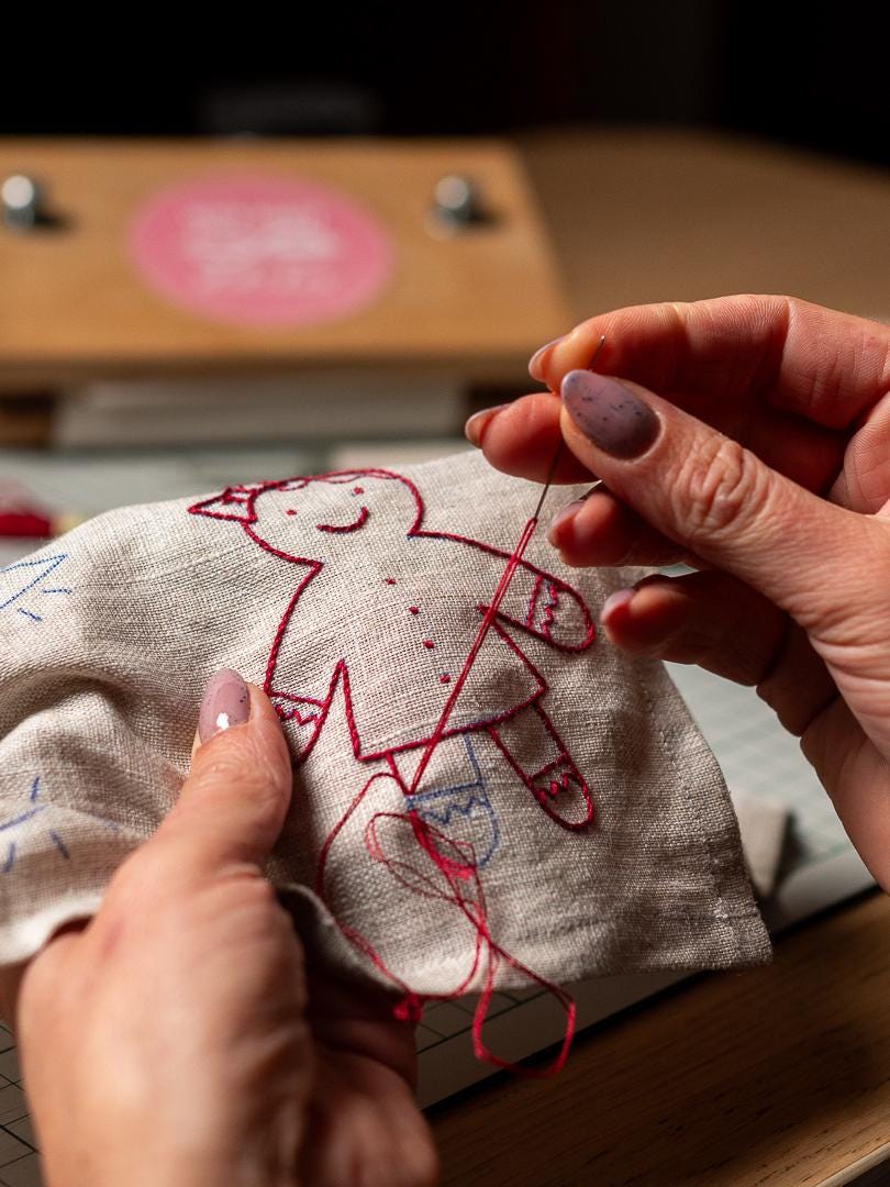 May include: Close-up of a hand-stitched gingerbread man design on a linen fabric. The embroidery uses red thread for the outline and details, with blue accents. The fabric is held in place, showing the process of hand embroidery.