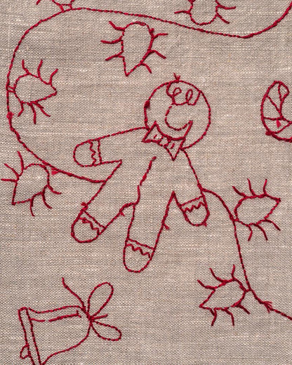 May include: Close-up of a beige fabric with red embroidery. The embroidery features a gingerbread man, a bell, and other festive shapes. The stitches are visible, creating a textured effect. The design is likely for a holiday-themed item.