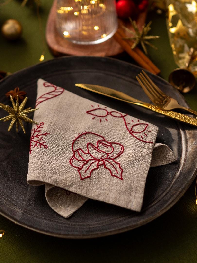 May include: A folded linen napkin with red Christmas-themed embroidery, including a candy cane, holly, and string lights, rests on a dark gray plate. Gold cutlery and decorative accents complete the festive table setting.