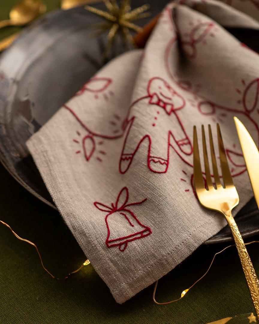 May include: A folded linen napkin with a red embroidered Christmas design featuring a gingerbread man, bell, and string lights. The napkin is placed on a dark plate with gold silverware, creating a festive table setting.