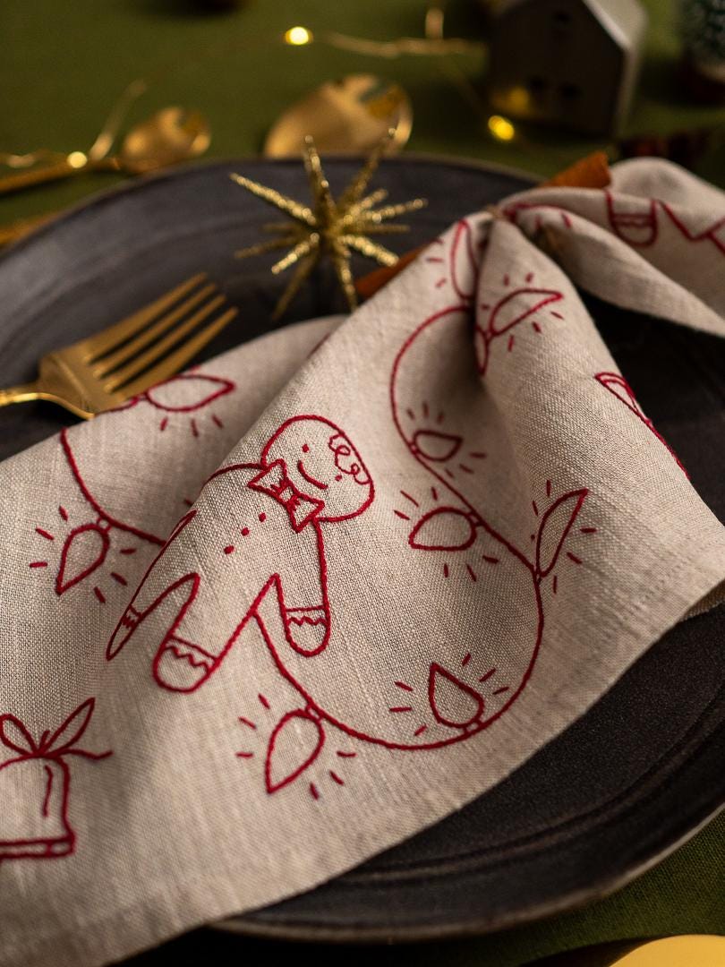 May include: A folded beige linen napkin with a red embroidered gingerbread man and string of lights design. The napkin is placed on a dark gray plate with a gold fork visible. Festive holiday table setting.