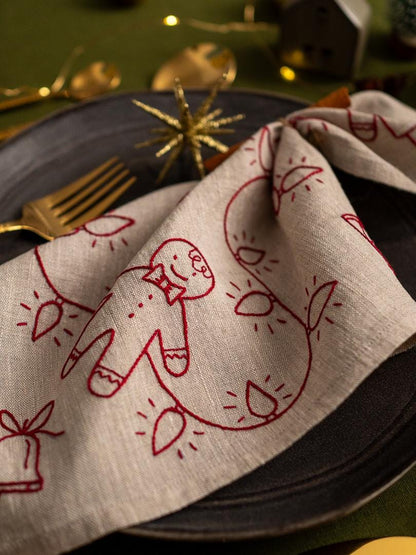 May include: A folded beige linen napkin with a red embroidered gingerbread man and string of lights design. The napkin is placed on a dark gray plate with a gold fork visible. Festive holiday table setting.