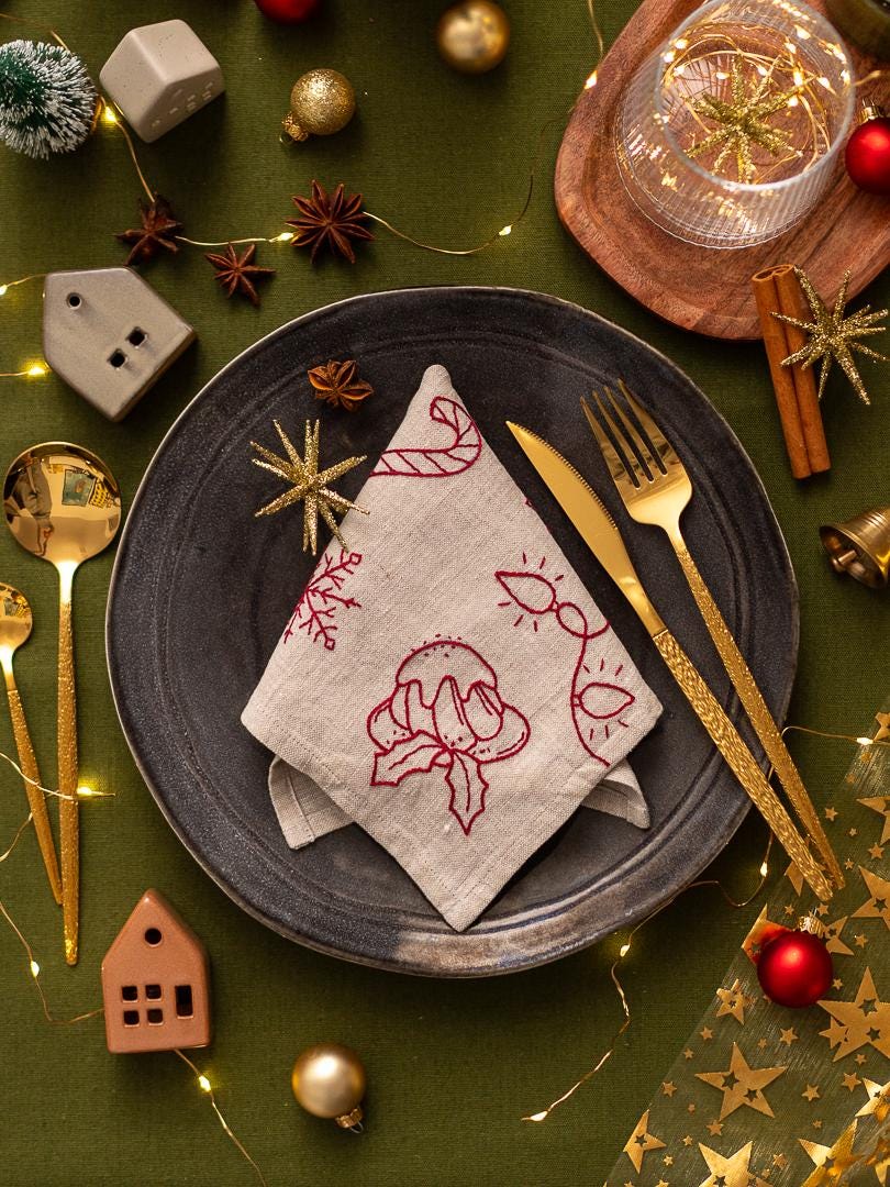 May include: A festive holiday table setting with a dark gray plate, gold utensils, and a linen napkin embroidered with red Christmas designs. The scene includes decorative ornaments, star anise, and fairy lights on a green tablecloth.
