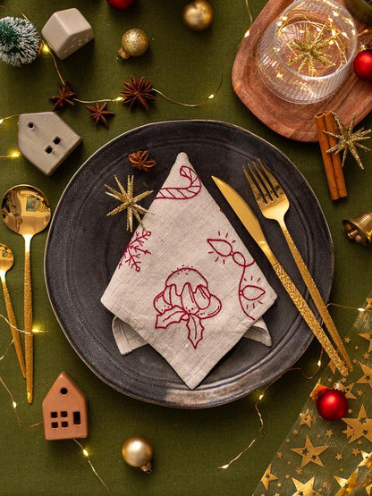 May include: A festive holiday table setting with a dark gray plate, gold utensils, and a linen napkin embroidered with red Christmas designs. The scene includes decorative ornaments, star anise, and fairy lights on a green tablecloth.