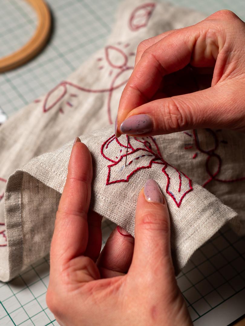 May include: Close-up of hand embroidery in progress. A beige linen fabric is being stitched with red thread, forming a floral design. The needle is being guided through the fabric, showcasing the detailed handwork. A wooden embroidery hoop is in the