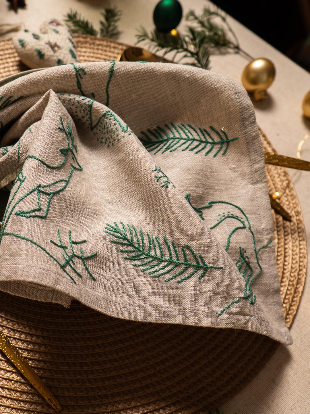 May include: Beige linen napkin with green embroidered Christmas tree and pine branch designs. The napkin is folded and placed on a woven placemat, with gold ornaments and greenery in the background. Festive holiday decor.