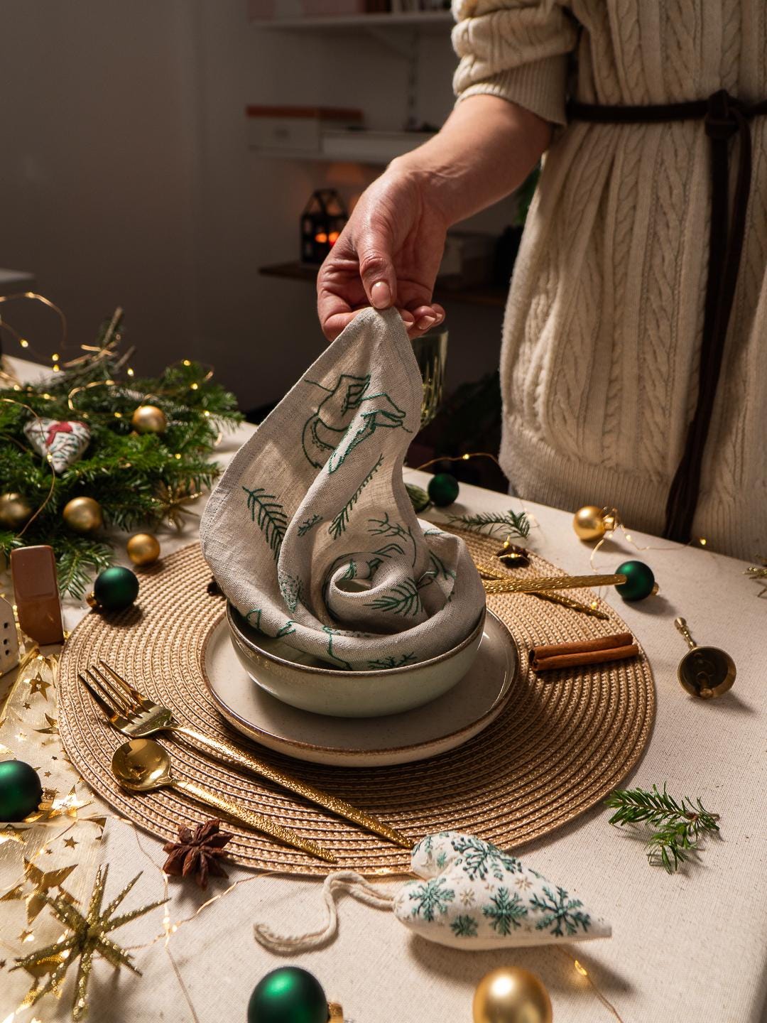 May include: A festive table setting with a folded linen napkin featuring a green tree design, placed in a bowl on a plate. Gold-colored flatware, green and gold ornaments, and a star-shaped decoration complete the holiday theme.