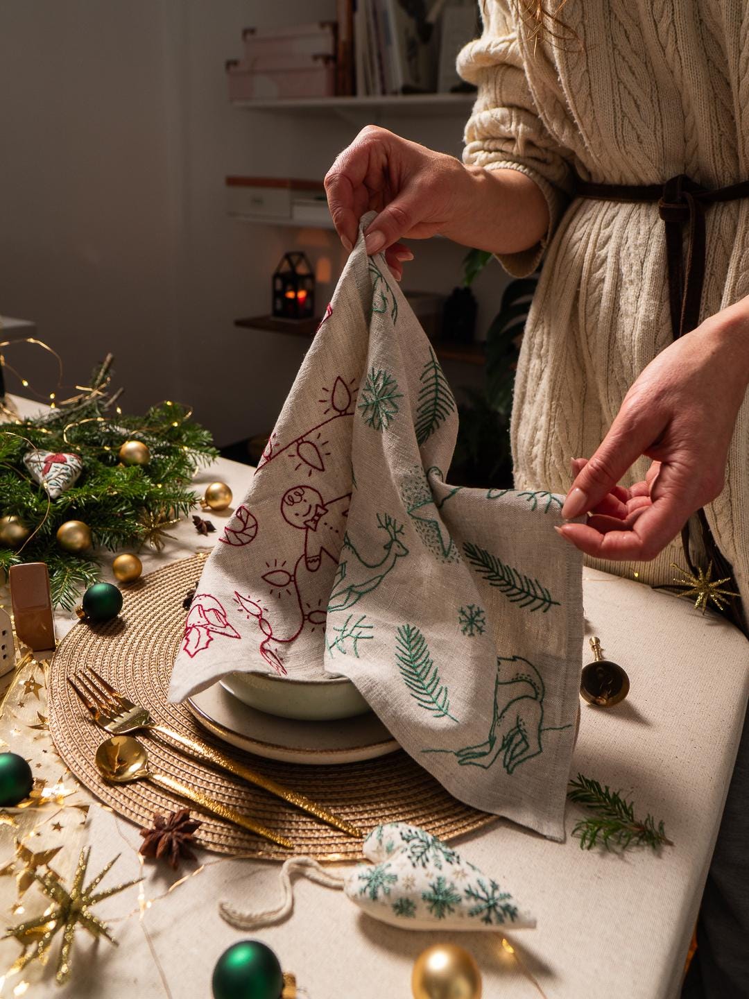 May include: A linen napkin with embroidered Christmas-themed designs in red and green, including a reindeer and festive lights. The napkin is being held over a plate, with gold cutlery and Christmas ornaments on the table.