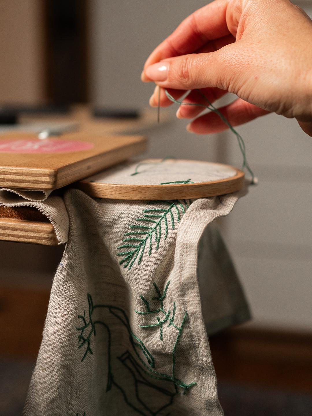 May include: Close-up of embroidery in progress. A hand holds a needle with green thread, stitching a design onto a linen fabric stretched within a wooden embroidery hoop. The fabric features green embroidered botanical elements.