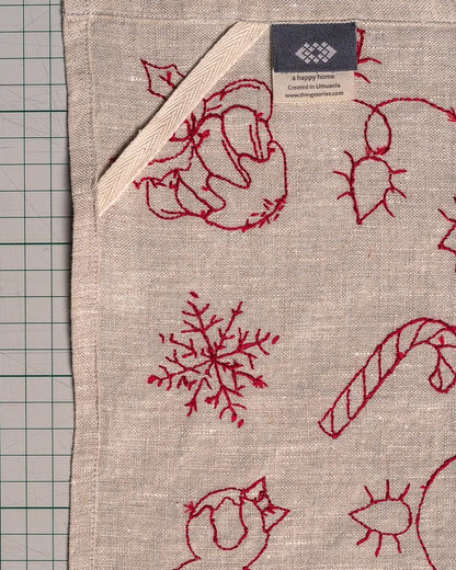 May include: A beige linen towel with red embroidered Christmas-themed designs, including a snowflake, candy cane, and floral motifs. A white ribbon loop is attached to the corner. The tag reads "a happy home Created in Lithuania".
