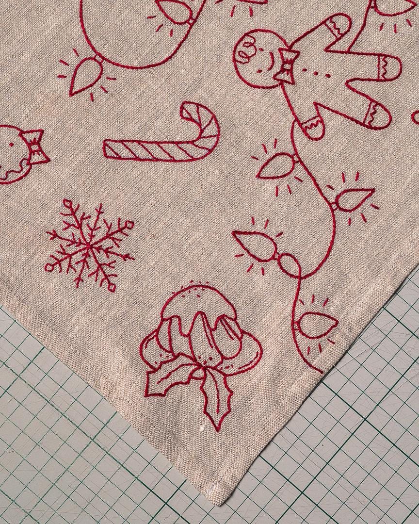 May include: Close-up of a beige fabric with red embroidered Christmas designs. The embroidery includes a gingerbread man, candy cane, snowflake, and Christmas lights. The fabric is likely a tablecloth or decorative piece.