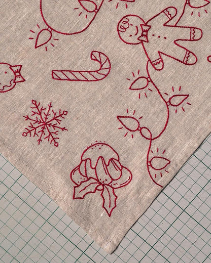 May include: Close-up of a beige fabric with red embroidered Christmas designs. The embroidery includes a gingerbread man, candy cane, snowflake, and Christmas lights. The fabric is likely a tablecloth or decorative piece.