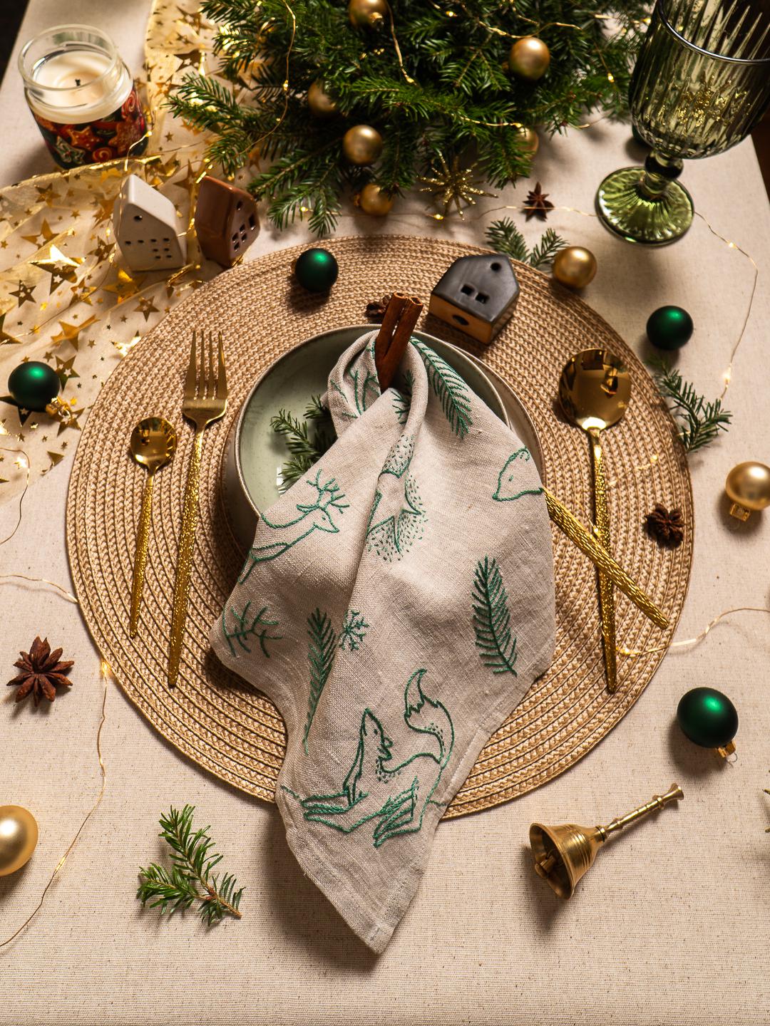 May include: A festive table setting with a woven placemat, gold cutlery, and a linen napkin embroidered with green foxes and pine branches. The scene includes green and gold ornaments, a candle, and miniature houses, creating a holiday atmosphere.