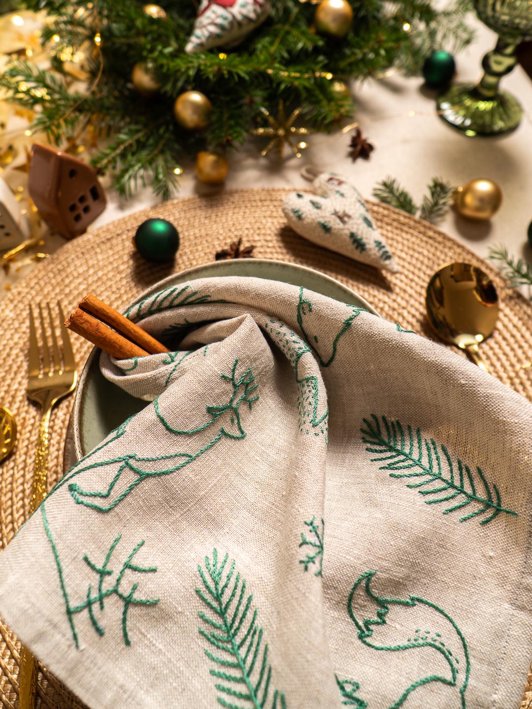 May include: A festive table setting with a linen napkin featuring green embroidered forest designs, including trees and a fox. Cinnamon sticks rest in a green bowl, with gold cutlery and Christmas ornaments in the background.