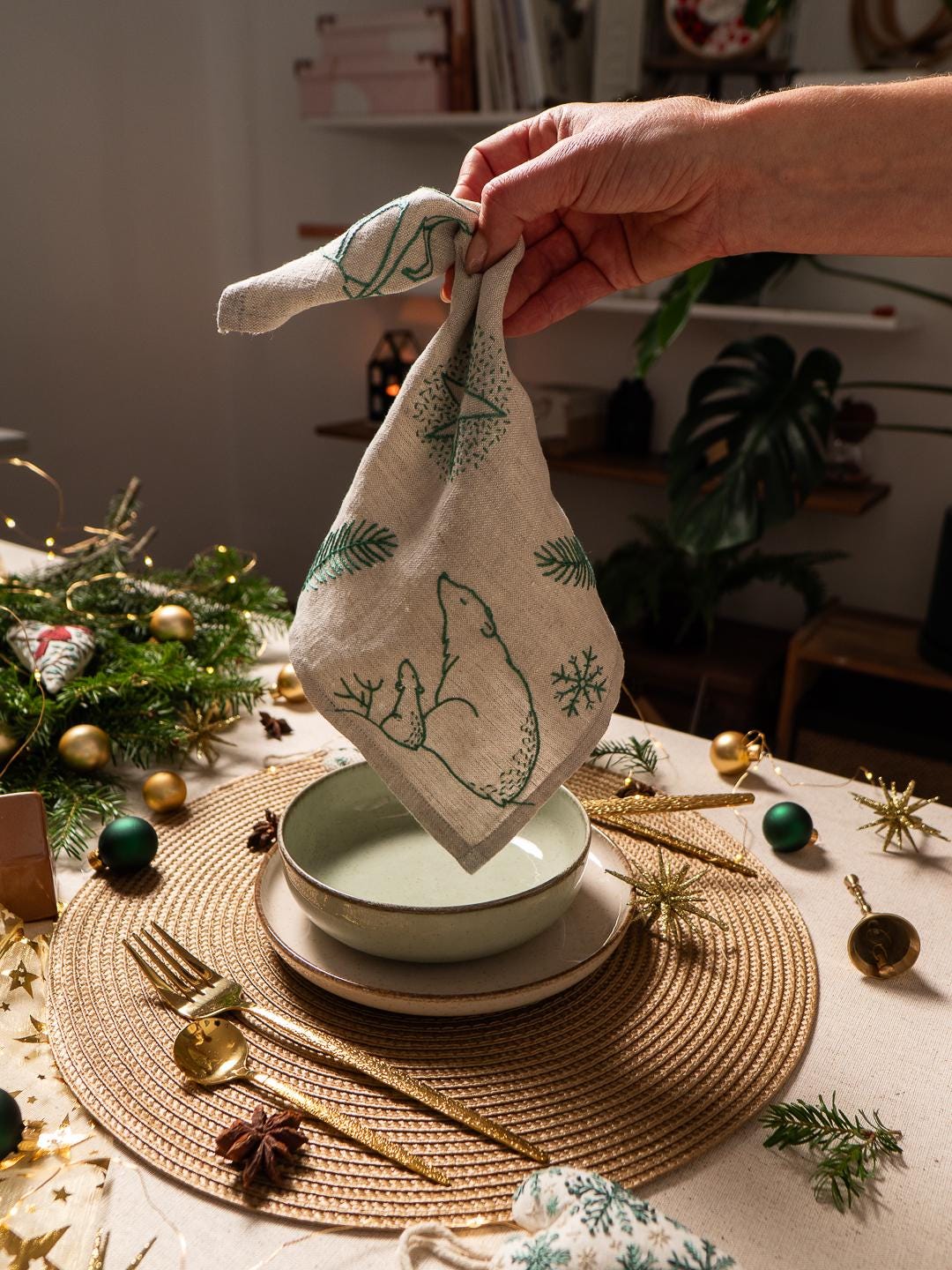 May include: A folded linen napkin with a green polar bear and snowflake design. The napkin is held above a table setting with a bowl, plates, and gold utensils. Christmas decorations are scattered around the table.