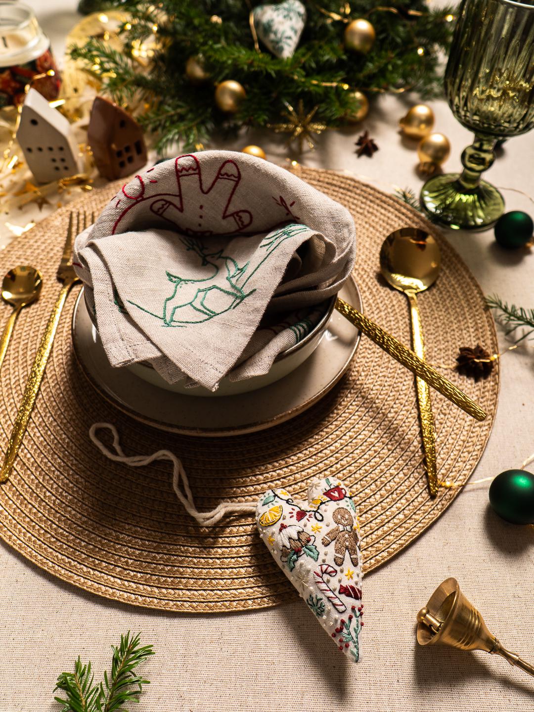 May include: A festive Christmas table setting with gold utensils, a woven placemat, and a stack of linen napkins embroidered with holiday designs. A heart-shaped ornament and a small bell add to the seasonal decor.